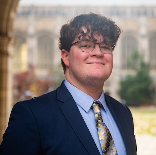 A photo of a young man in glasses. He is standing in the UM-Ann Arbor law quad outdoor area. He's wearing a navy blue suit, a light blue dress shirt and a tie