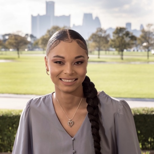 Portrait of a young smiling woman wearing a gray dress and a heart necklace. There is a city landscape in the background