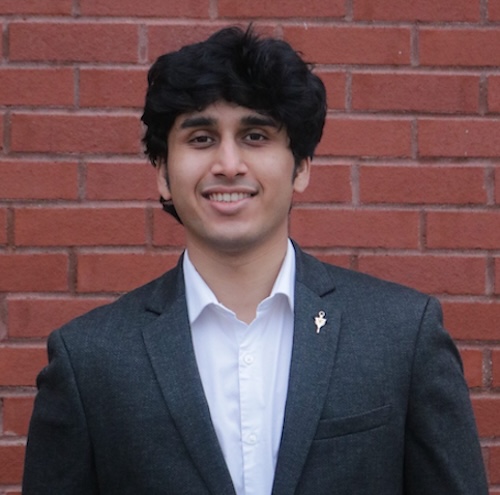 A male student with short black hair; he is wearing a gray suit and a light blue dress shirt