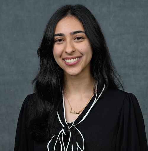 A portrait of a smiling woman with long black hair who is wearing a black blouse with a black and white scarf around her neck. The background of the photo is blue