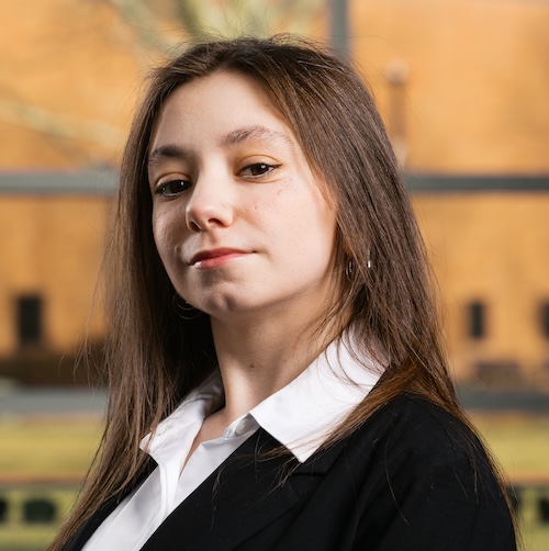 Photo of a young woman with long brown hair. She has a slight smile and is wearing a white dress shirt and a black suit jacket