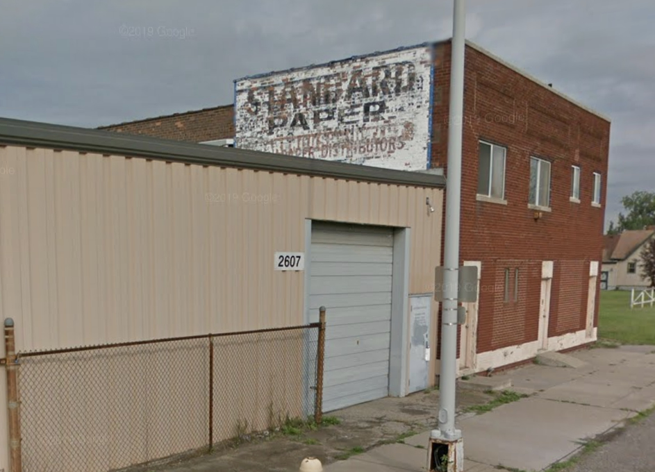 An old building with red brick with a white painted sign that has black letters. It reads, Standard Paper Company.