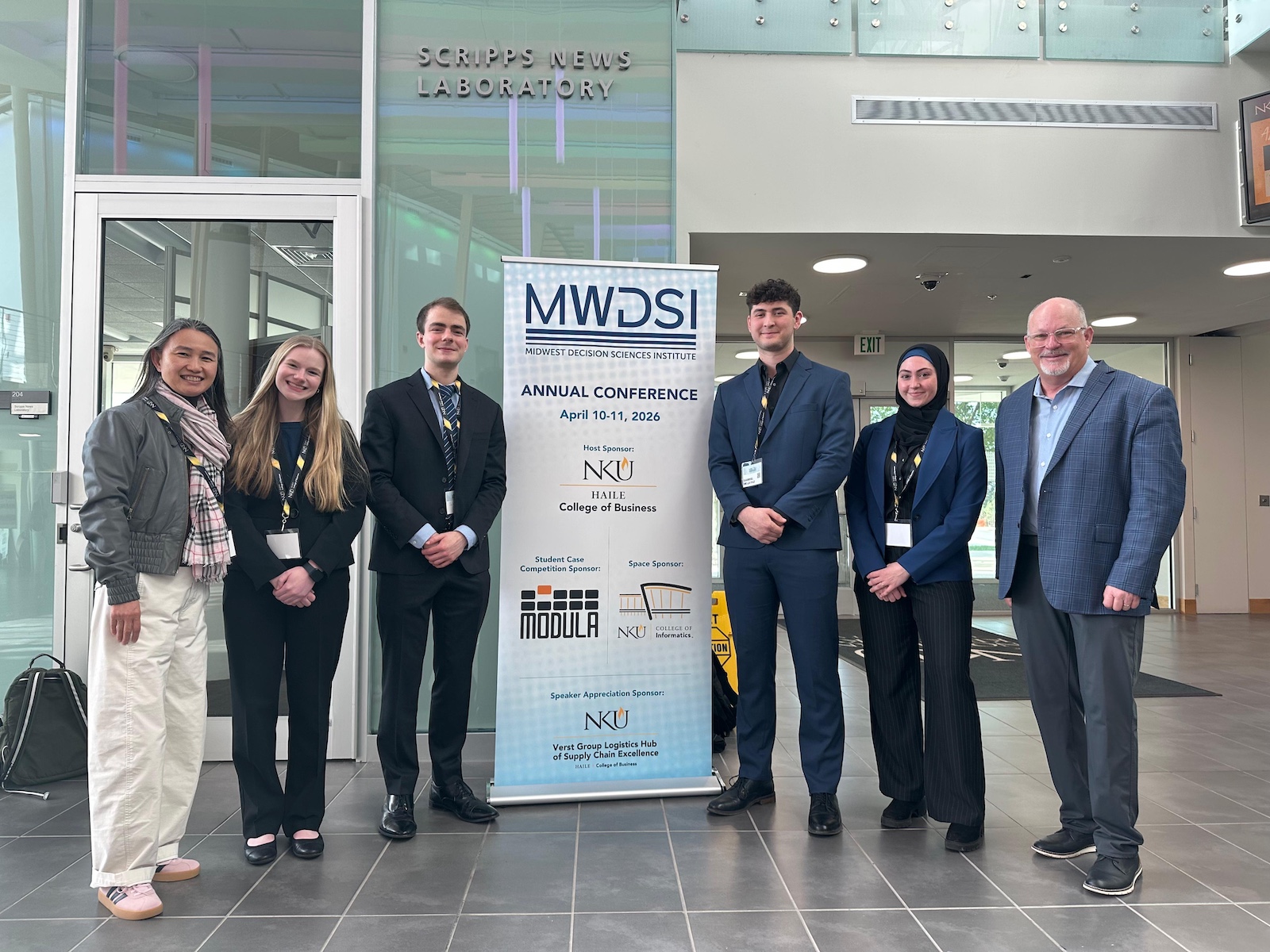 Group of six people in business attire standing in a university lobby beside a banner for the Midwest Decision Sciences Institute Annual Conference (April 10–11, 2026) at Northern Kentucky University’s Haile College of Business; “Scripps News Laboratory” signage visible in the background.