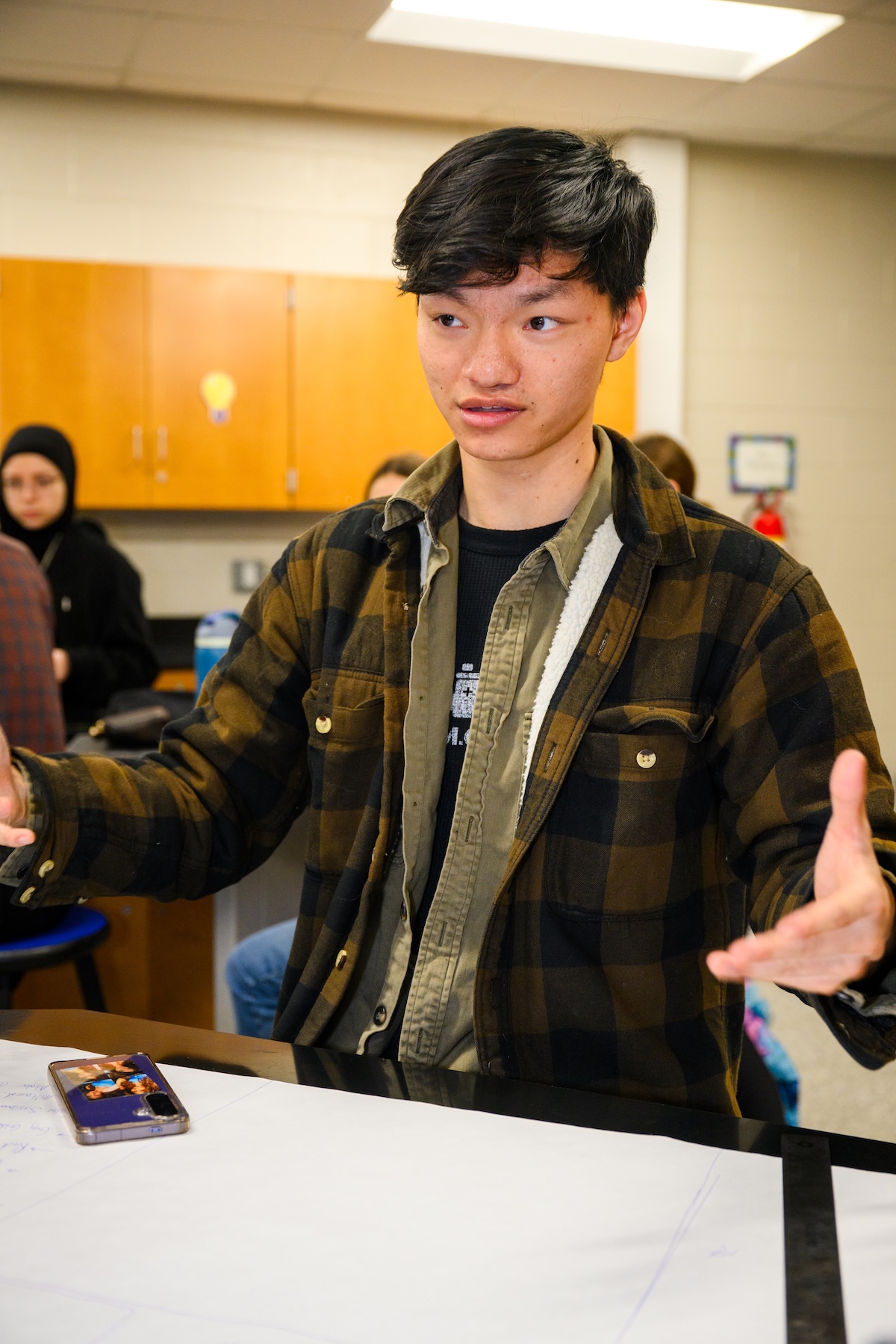 A male sits at a table and has his hands extended in a gesture while speaking
