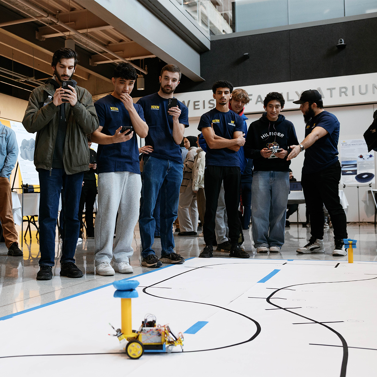 A group of individuals observes a small robot on the floor in a modern indoor space. Some are holding and using smartphones. The setting appears to be a demonstration or competition area with visible pathways marked on the floor.