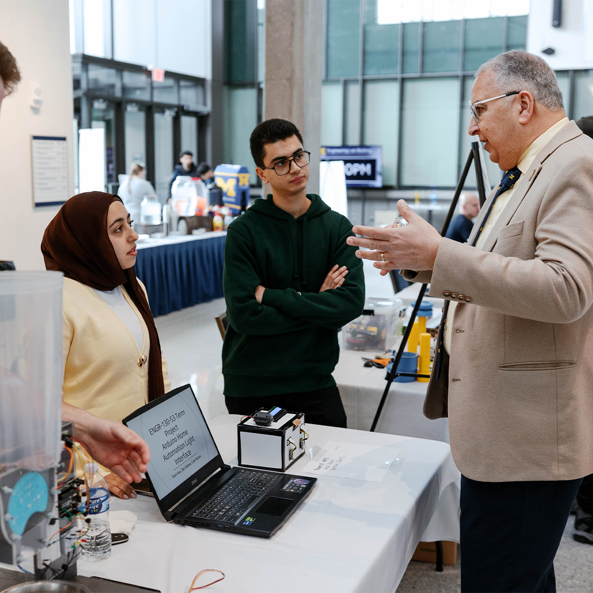 A person in a beige suit speaks to two individuals at a technology exhibit. A table displays a laptop and some tech devices. 