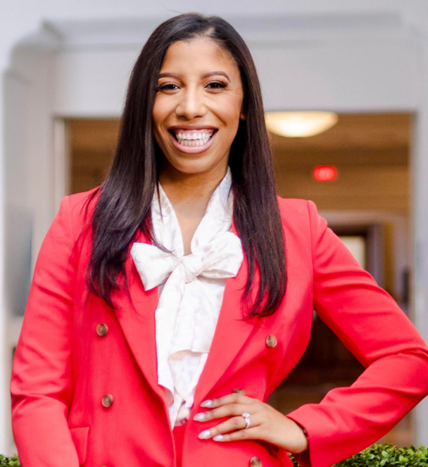 Portrait of a person with long dark hair wearing a bright pink blazer.They have a white bow blouse and one hand posed on their hip.Indoor hallway background is softly blurred, with the face obscured for privacy.
