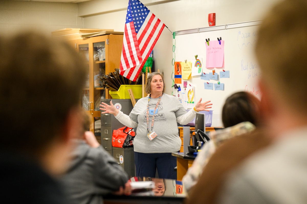 A female teacher speak to a classroom of students. There is a white board and an American flag in the background