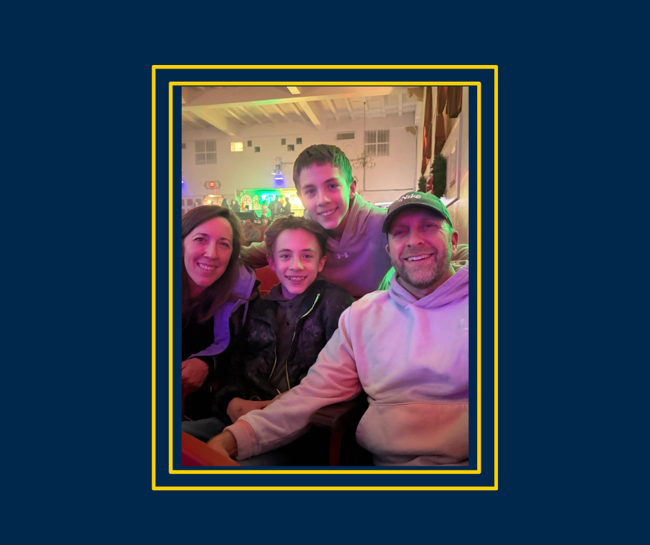 Four people—two adults and two children—smiling and sitting together indoors at what appears to be a lively venue or event space with colorful lighting in the background.