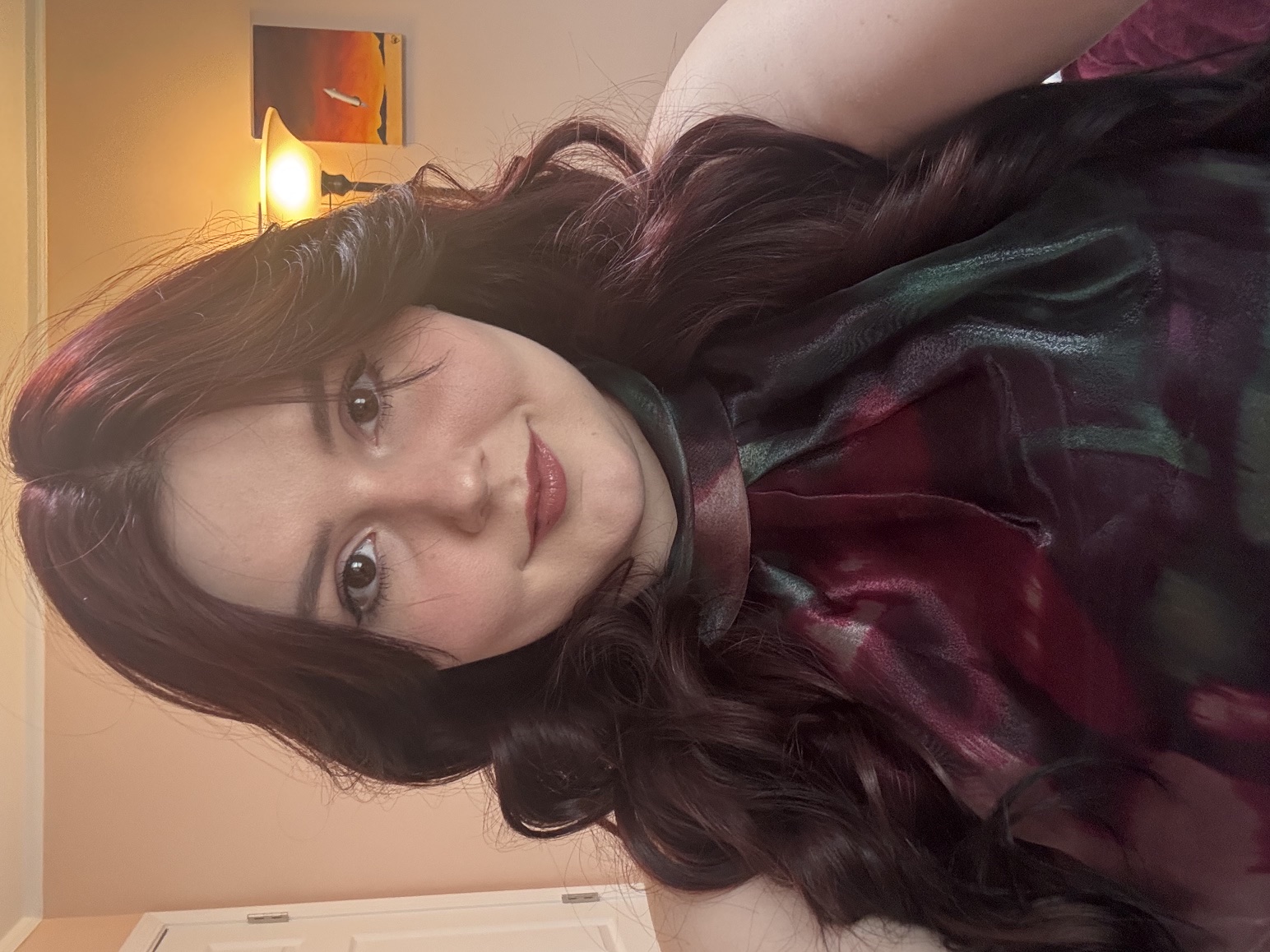 Person with long, wavy dark burgundy hair posing indoors.Face is obscured; wearing a sleeveless burgundy and green patterned top.Warm bedroom background with a lamp, wall art, and a white door.