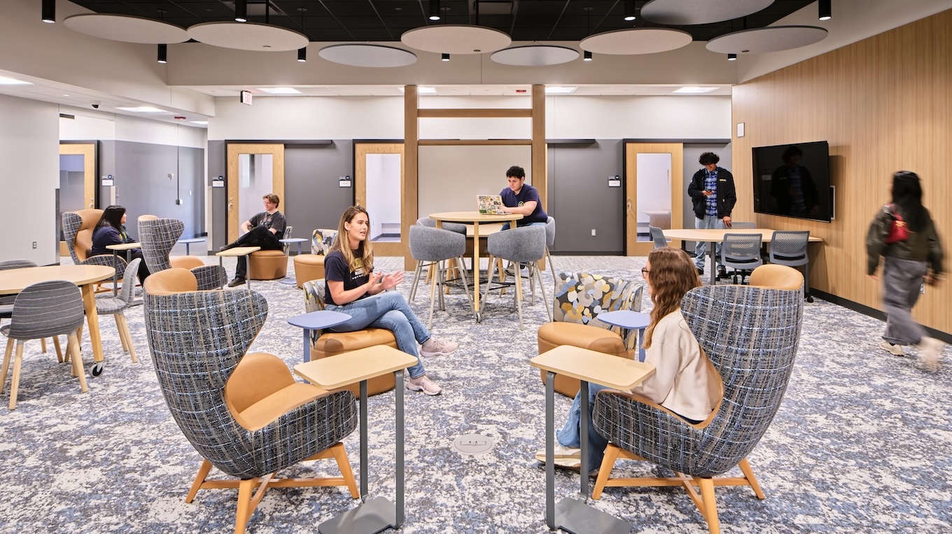 People sit chairs and at tables in an open lobby area. There are tv monitors on the wall and a black ceiling that has round lights.