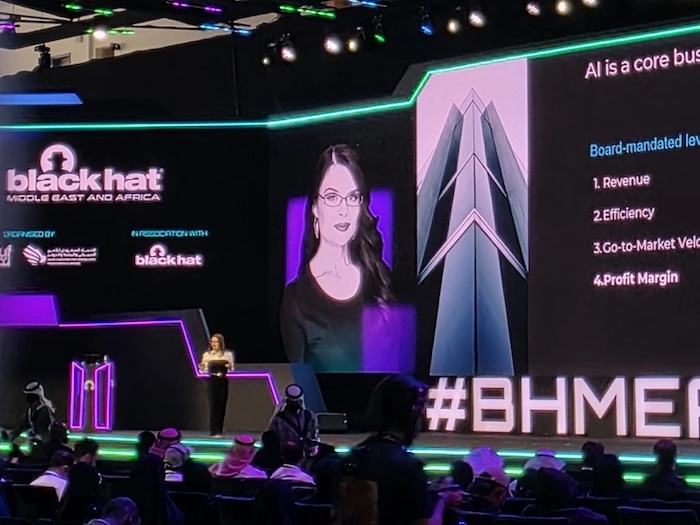 A woman speaks in front of an audience. She's on a large stage with a portrait of herself in the background. 