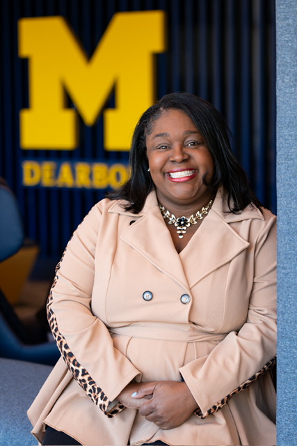 Person seated indoors wearing a light tan coat and necklace.Blurred University of Michigan “M” and Dearborn sign in the background.Portrait-style campus image with the person centered in frame.