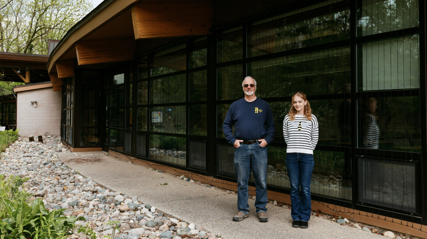 Two people stand on a paved walkway outside a modern building with large dark-framed windows, a curved wooden roof overhang, and landscaped rocks and greenery.