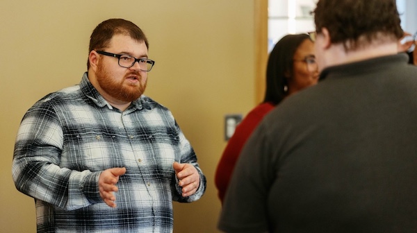 A man wearing glasses and a gray plaid shirt speaks with his hands raised, facing two other people during an indoor conversation.