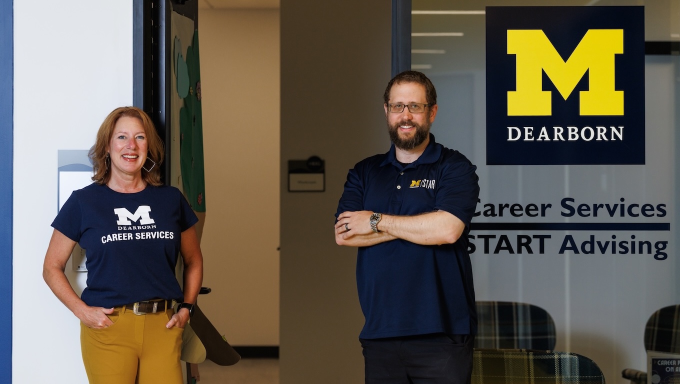 Two smiling UM-Dearborn staff members stand outside of an office entrance. On the right, a glass wall features a large yellow UM-Dearborn Block M, along with the text "Career Services" and "START Advising."
