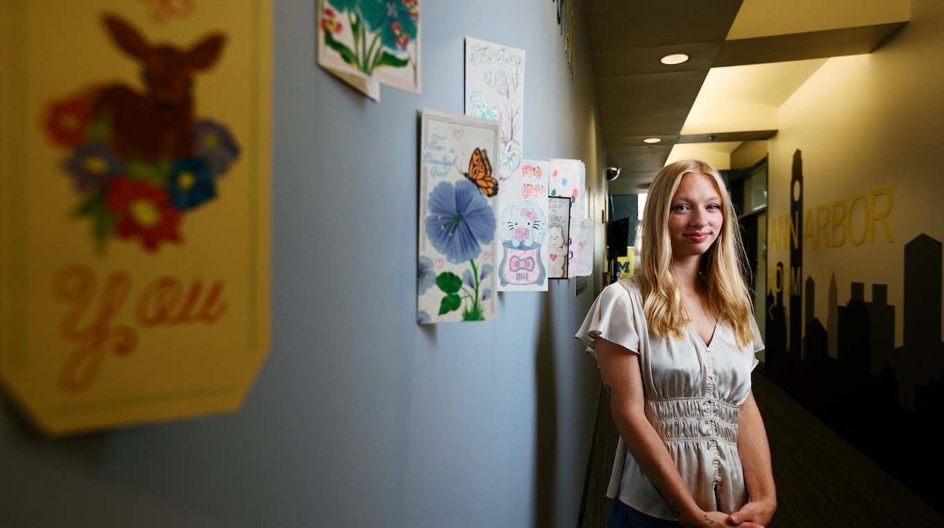 A young woman with long blonde hair and a light cream-colored blouse stands smiling in a hallway. On the wall to her left, several colorful greeting cards and artwork featuring flowers, a deer, and Hello Kitty are displayed in a line.