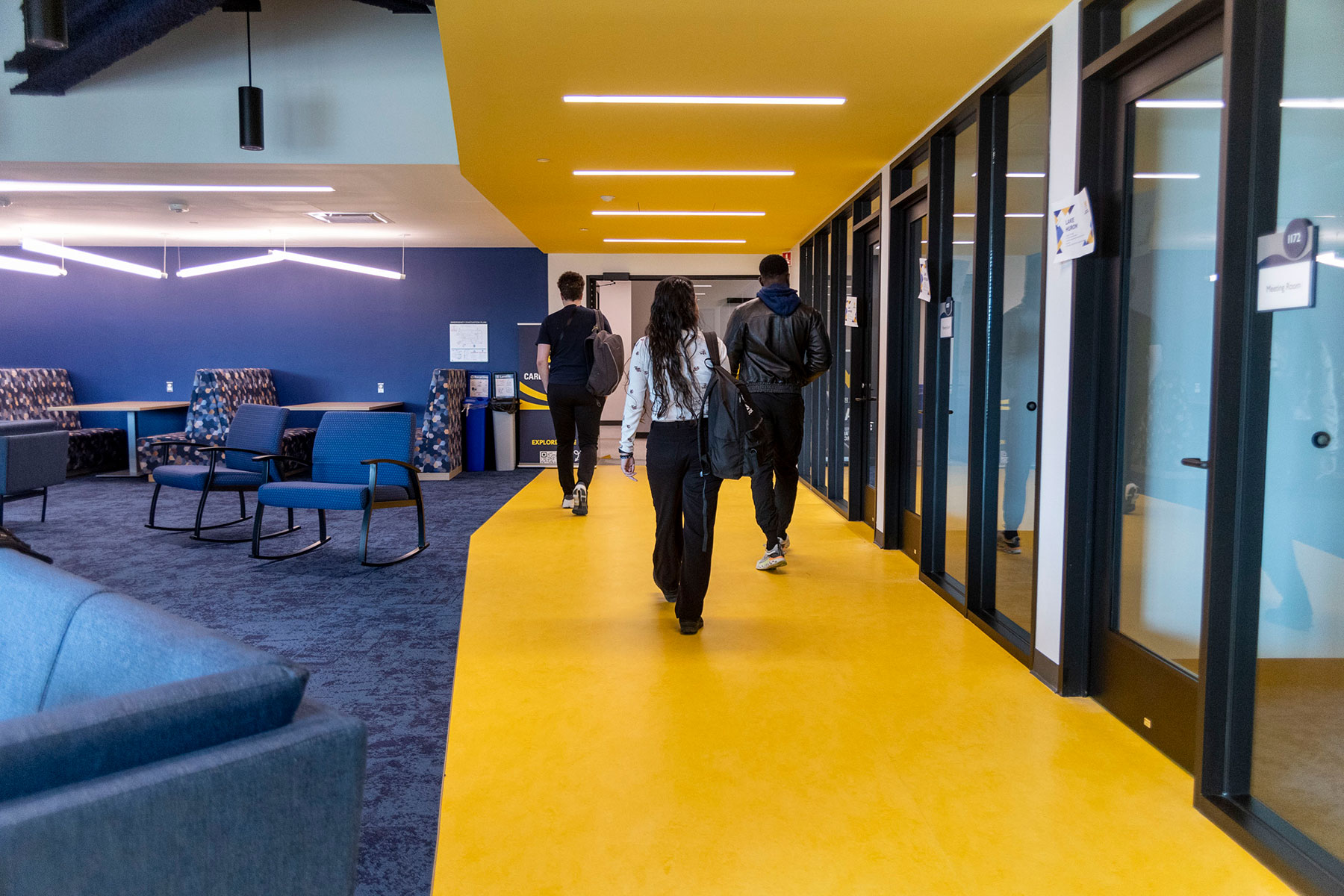 Three students walk through a modern campus interior with a bright yellow floor and ceiling, glass-walled meeting rooms, blue lounge seating, and study areas.