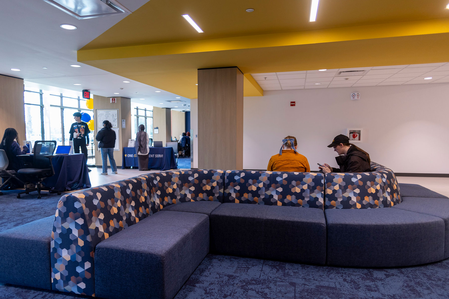 Students sit and gather in a modern campus lounge with curved blue patterned seating, blue carpet, large windows, service tables, and a yellow ceiling feature.