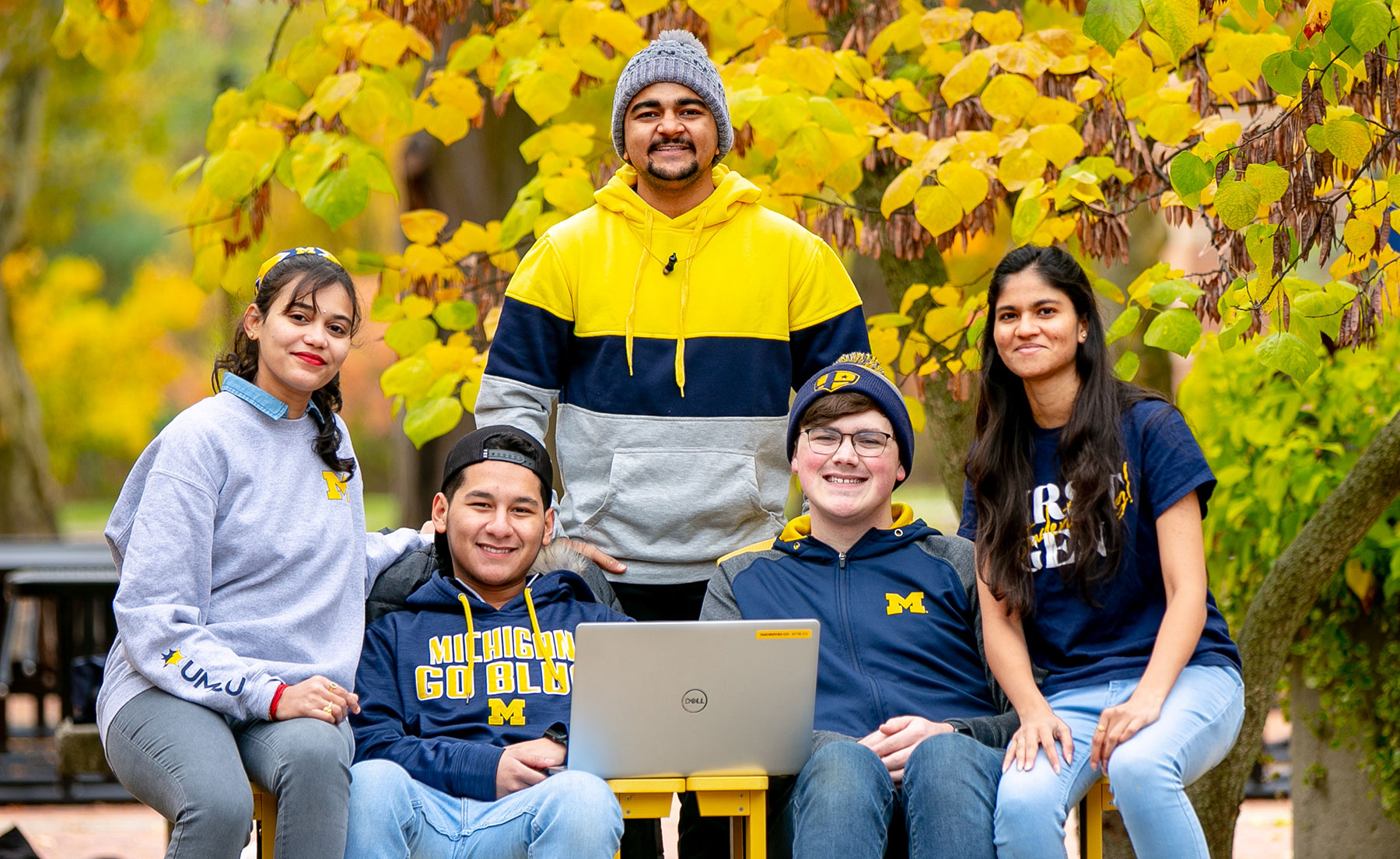 Students look at the camera while studying on a laptop near Chancellor’s Pond on a fall day. They are wearing University of Michigan apparel.
