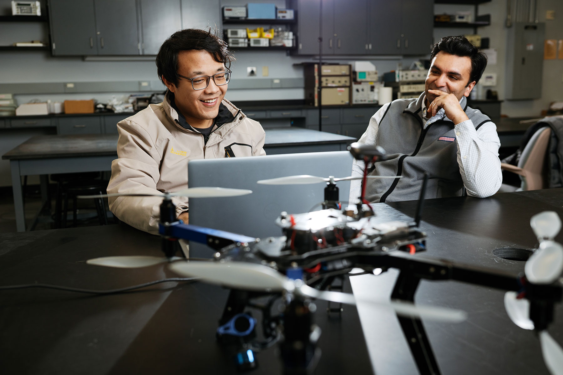 Two male University of Michigan students sitting at a lab desk looking at a laptop. One has glasses and a tan jacket, and the other is in a dress shirt with a fleece vest.