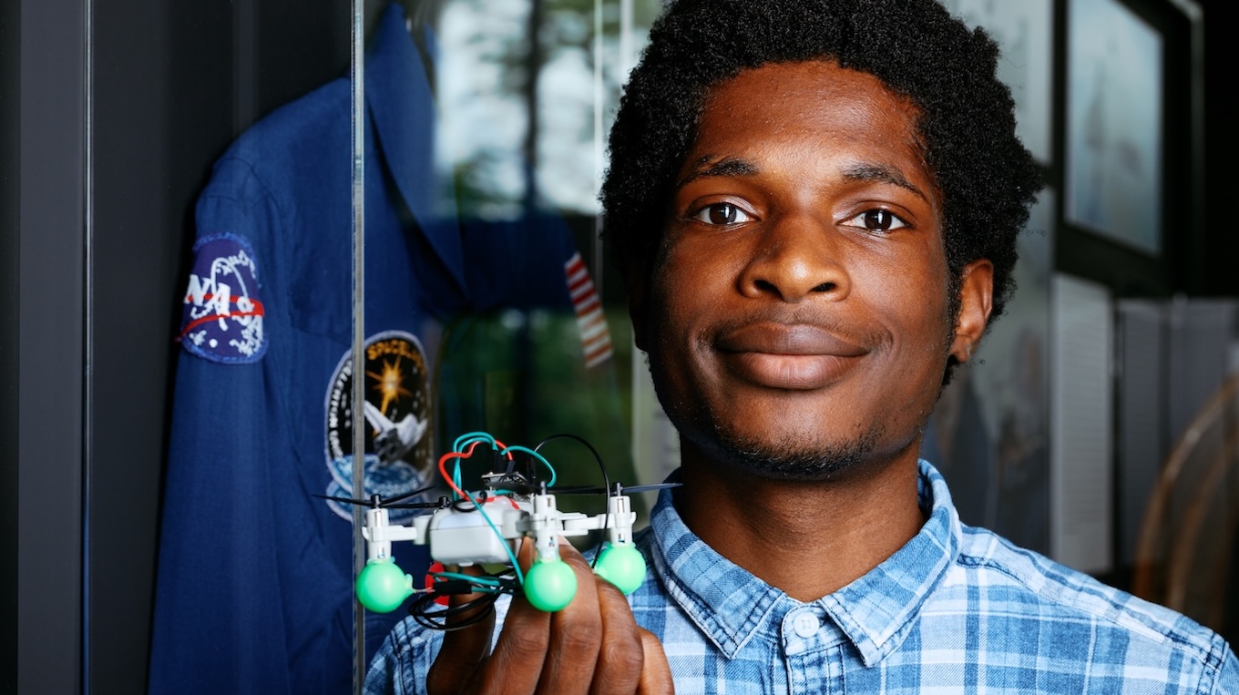Close-up photo of a young Black man wearing a blue plaid button-down shirt, holding a small custom quadcopter drone with exposed wiring and green padded feet. He smiles slightly at the camera. In the background, a blue NASA flight jacket with official patches is displayed inside a glass case.