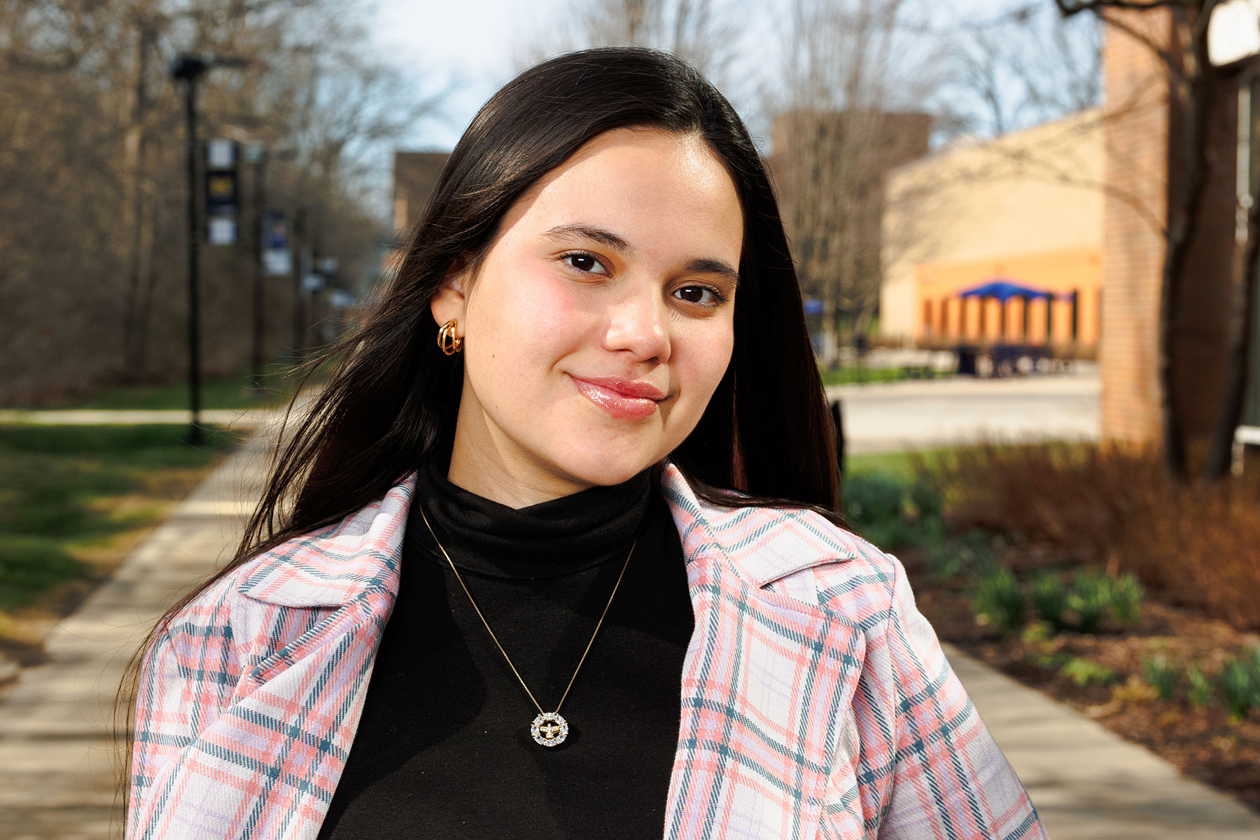Young woman standing outdoors on a campus walkway, wearing a pink plaid jacket and black top.