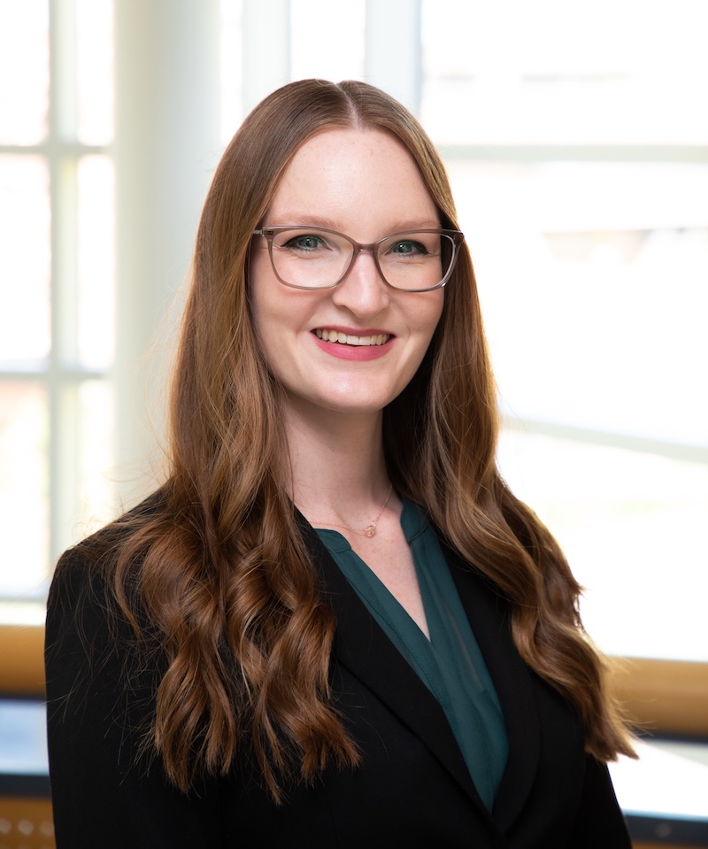 Professional headshot of a person with long wavy auburn hair.They wear a black blazer over a teal blouse and a small necklace.Bright indoor background with large windows softly out of focus.