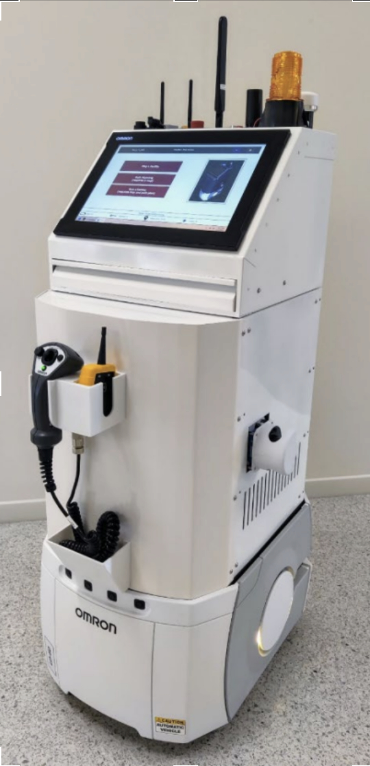 A white Omron autonomous mobile robot equipped with a touchscreen display, safety light, antennas, and side-mounted controllers standing indoors on a tiled floor.