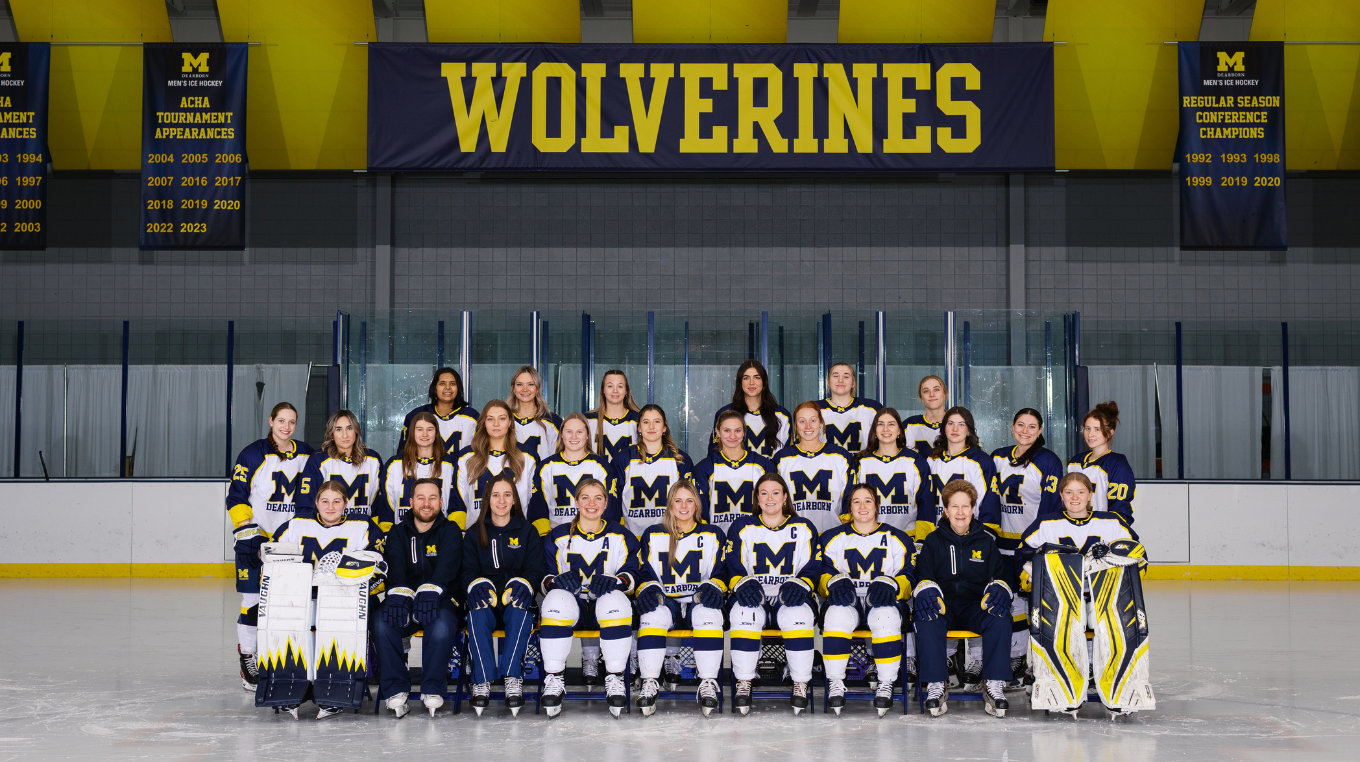 University of Michigan-Dearborn women’s ice hockey team poses for a group photo on the ice in uniform, with coaches seated in front and a large “WOLVERINES” banner hanging above them.
