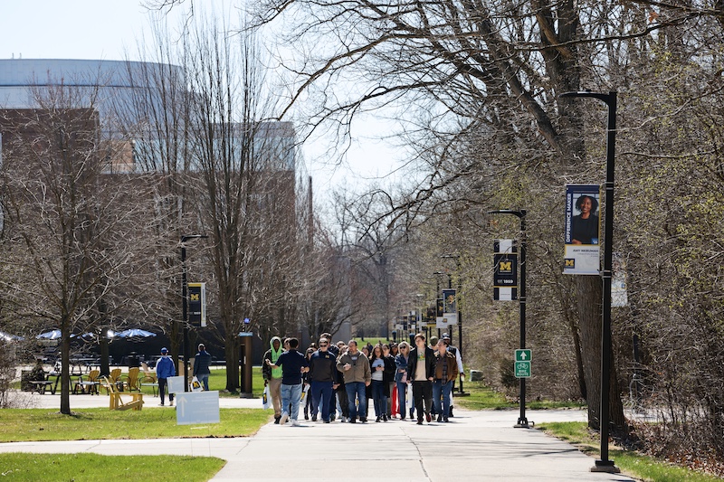 A group of people walks together on a wide sidewalk through a University of Michigan campus area. Leafless trees, lampposts, grass, and maize-and-blue banners line the path on a sunny day.