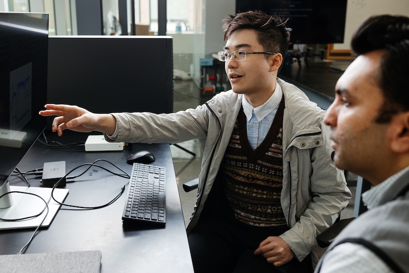 Two people collaborate at a computer workstation in a lab or office setting. One person wearing glasses points toward a monitor while the other looks on, suggesting a discussion about something on the screen.