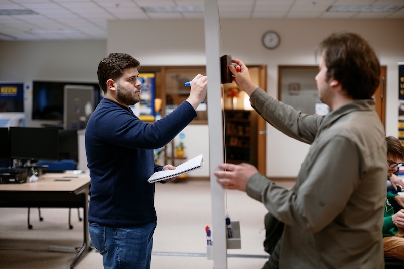 Two people stand on opposite sides of a clear whiteboard, writing with markers during a classroom or collaborative work session. One holds a notebook while computer workstations and classroom furniture appear in the background.