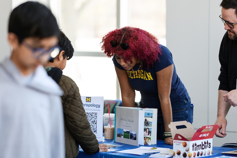 A person in a Michigan shirt leans over a blue table to speak with attendees at a campus display. The table includes an anthropology sign with a QR code, a tri-fold presentation board, papers, drinks, and a box of Timbits.