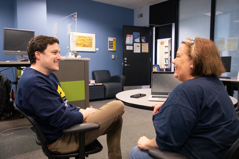 Two people in navy sweatshirts sit facing each other in a campus office, smiling during a conversation. A laptop and phone rest on a round table between them.