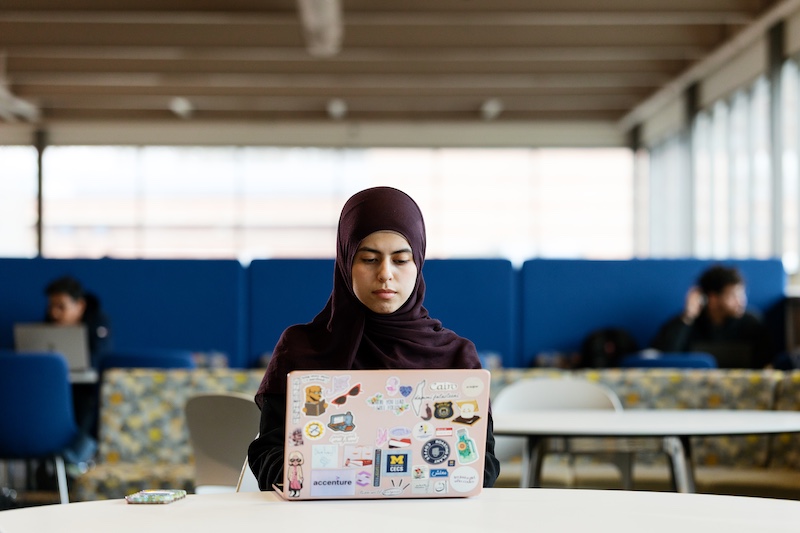 A student in a dark hijab sits at a table using a laptop covered with stickers, including a University of Michigan block M. Other students work in the background of a bright lounge with blue seating and large windows.