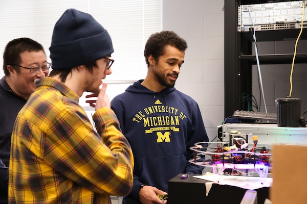 Three students gather in a lab around a small drone with glowing lights, with one wearing a University of Michigan hoodie.
