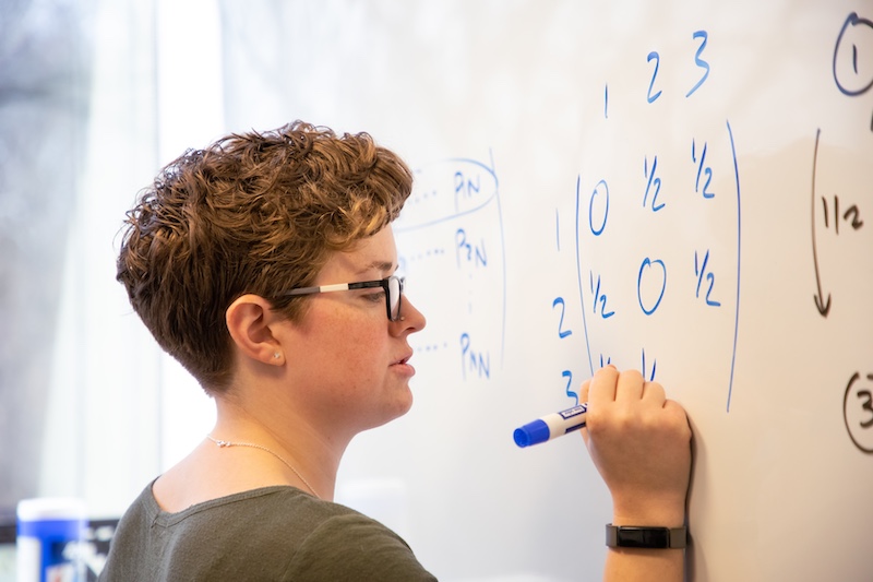 A person with short curly hair and glasses writes equations on a whiteboard, including a blue three-by-three matrix with entries such as 0 and 1/2.