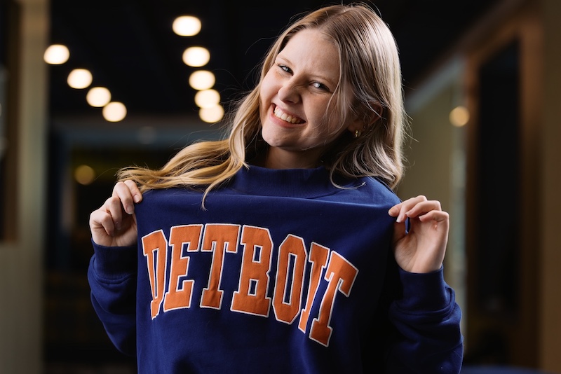 Young woman with long blonde hair smiles while holding the collar of a navy sweatshirt that says “Detroit” in large orange letters, standing in a softly lit indoor hallway.
