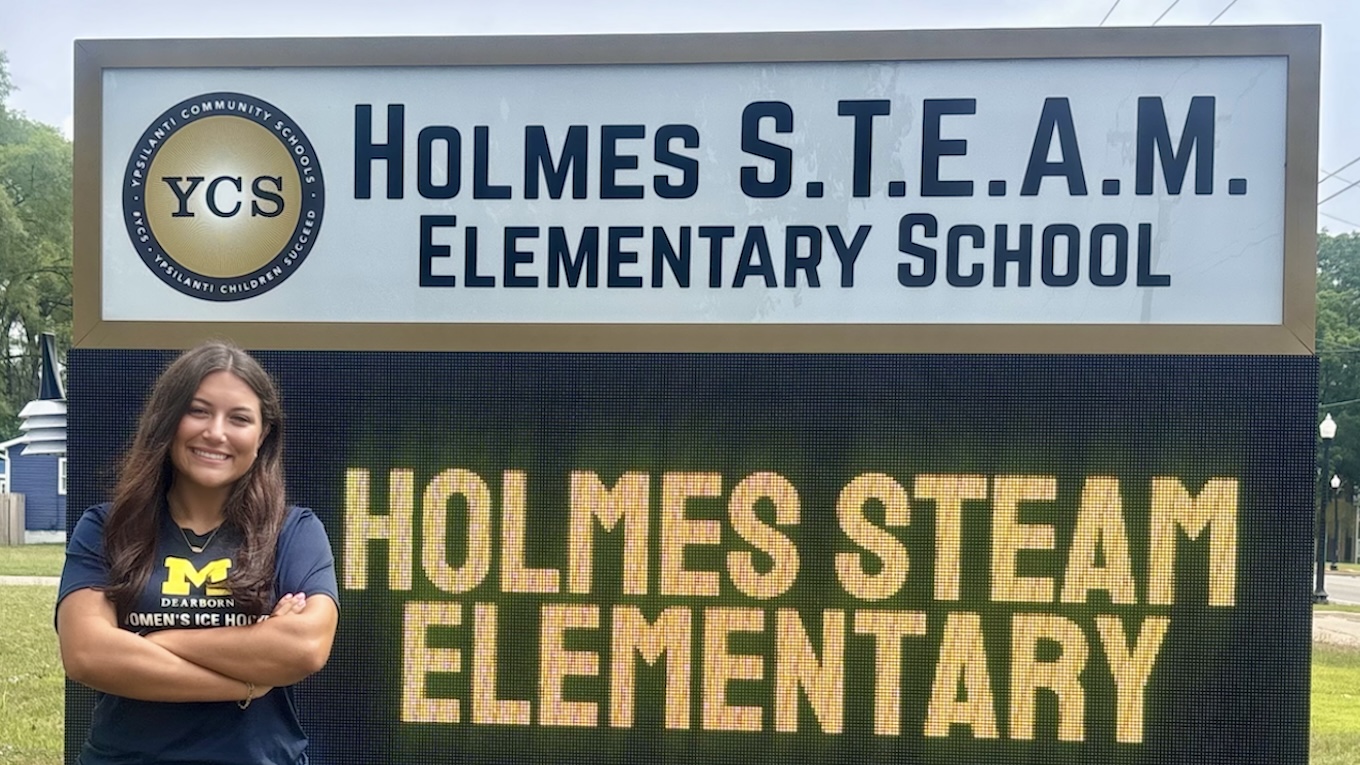 A smiling woman standing with crossed arms in front of an outdoor school marquee sign for Holmes S.T.E.A.M. Elementary School.