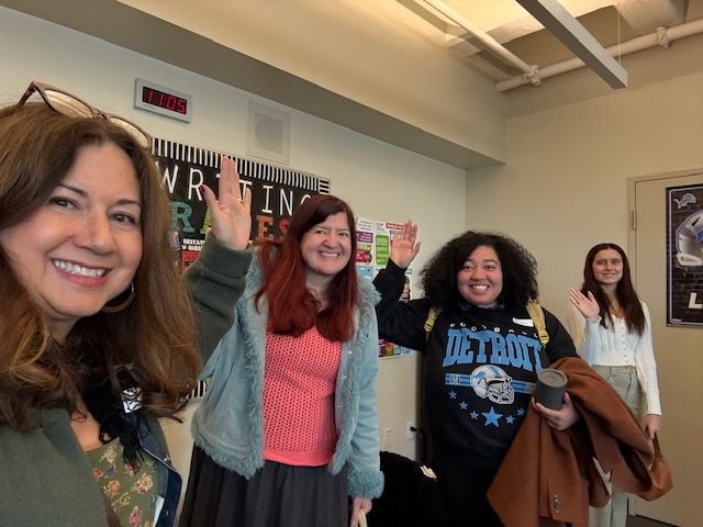 Four smiling women standing in a classroom lined up from front to back, all raising a hand to wave at the camera. In the background, a digital clock shows 11:05 and a poster reads "WRITING."