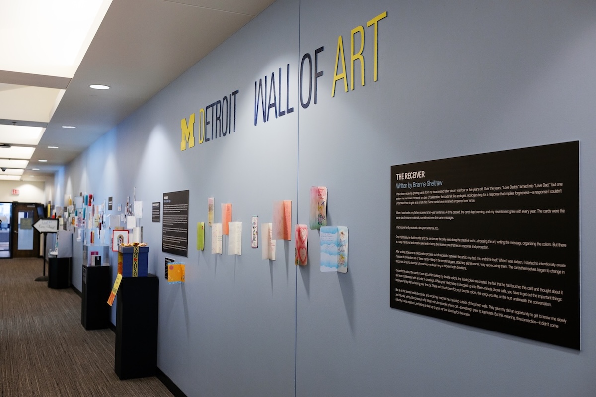 An indoor art exhibition hallway featuring a light blue wall labeled "M DETROIT WALL OF ART" displaying greeting cards and black informational plaques.