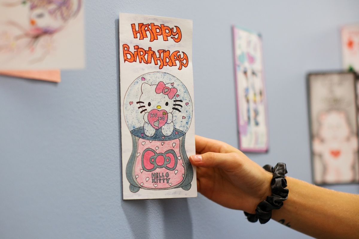 A hand wearing a black scrunchie places a homemade rectangular card onto a light blue wall. The card reads "HAPPY BIRTHDAY" at the top in orange bubble letters and features a drawing of Hello Kitty inside a snow globe holding a pink heart that says "XOXO".