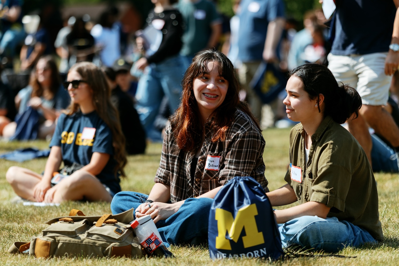Two female students sitting cross-legged on a sunny lawn during an outdoor campus event, chatting with each other near a blue UM-Dearborn drawstring bag.