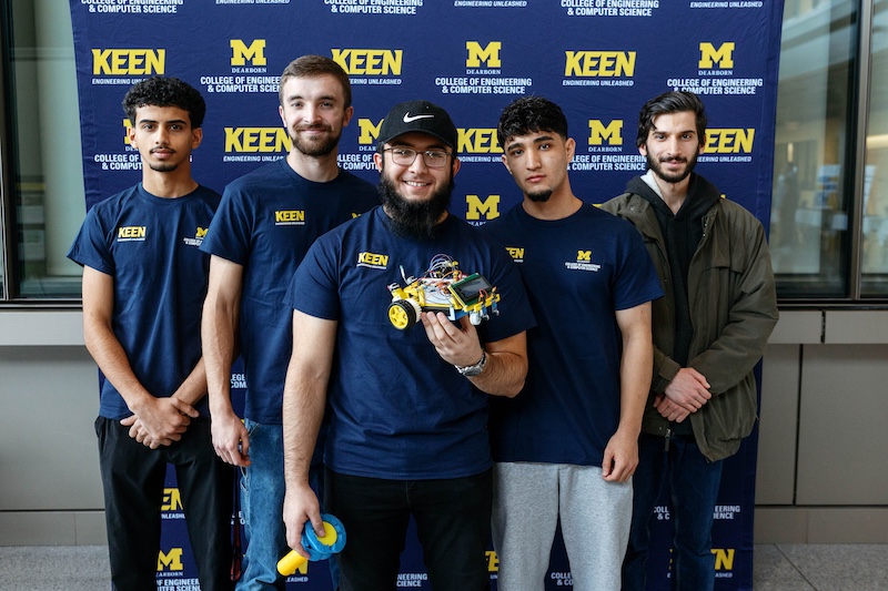 Five students stand together in front of a navy UM-Dearborn College of Engineering and Computer Science KEEN backdrop. Four wear navy KEEN shirts, and the student in the center holds a small wheeled robot with visible wiring and yellow wheels.