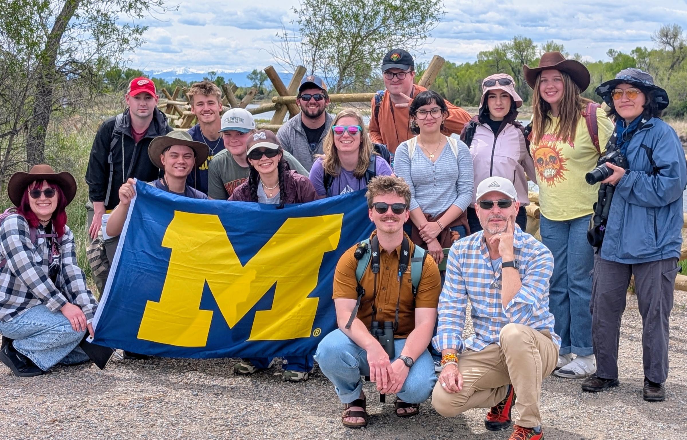 A group of UM-Dearborn students and faculty pose outdoors during a field experience, with several students holding a large maize and blue University of Michigan flag. The group is dressed in hiking and outdoor gear, with trees and mountains visible in the background.