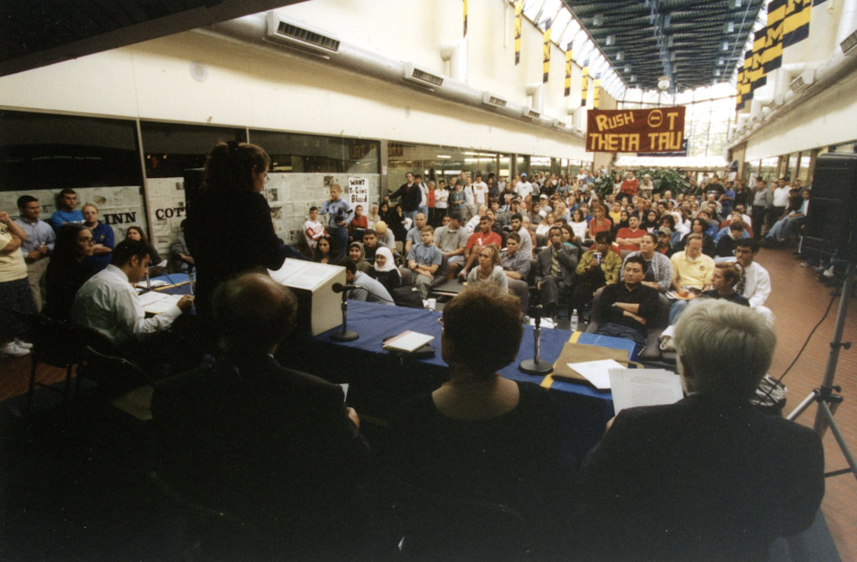 A speaker addresses a large crowd seated in a spacious indoor campus hallway during a panel discussion or town hall meeting.