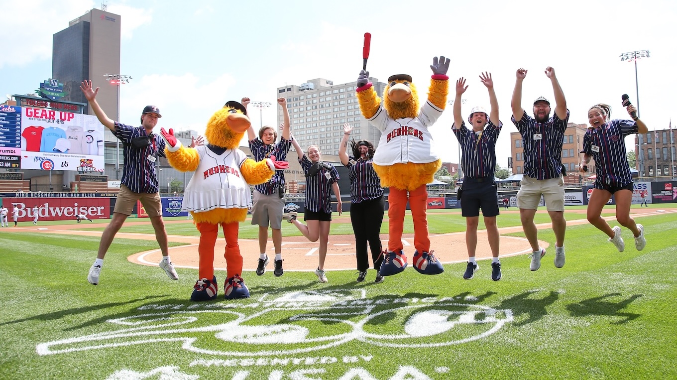 A group of seven energetic stadium staff members in matching pinstriped shirts leap and cheer on a baseball field alongside two yellow bird mascots wearing "Mud Hens" jerseys. The group is captured mid-air with arms raised on the green outfield grass, with the infield dirt, stadium seats, and city skyline visible under a bright, sunny sky.