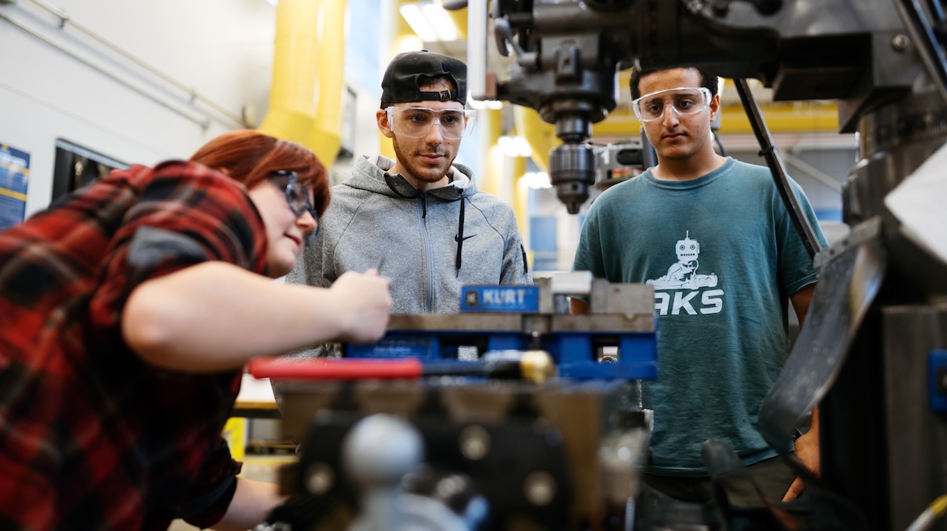 Three engineering students wearing protective safety glasses gather around a milling machine in a workshop.