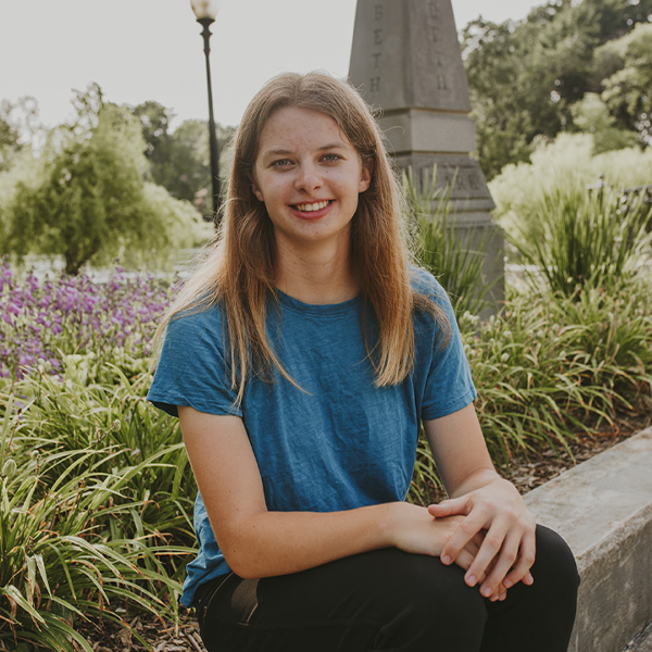 Person with long blonde hair sitting on a bench in a park with a blue t-shirt.